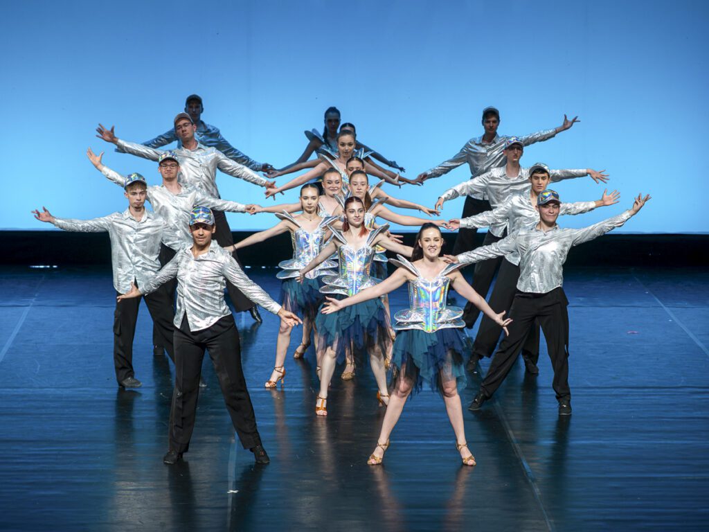 Kluboví tanečníci tanečného športu K1 Tanečnej školy NEXUM v skupinovej choreografii, v strieborných kostýmoch na javisku s modrým osvetlením